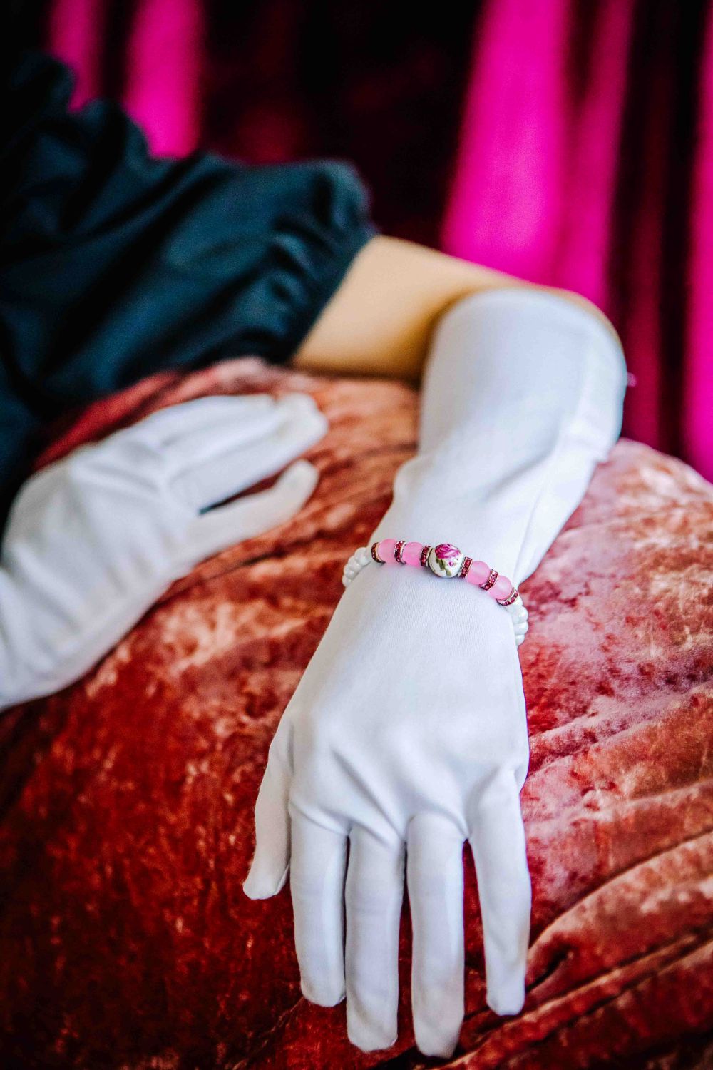 Person wearing white gloves with a pink beaded bracelet on a red cushion against a pink curtain background