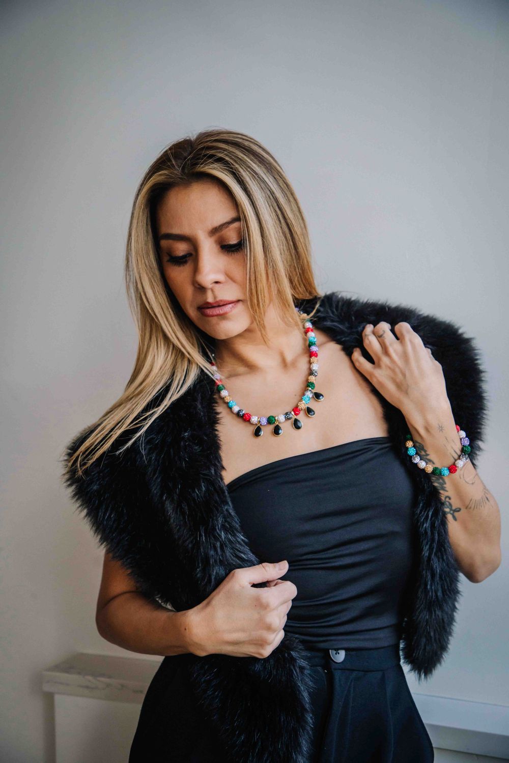 Woman wearing a black fur vest and colorful necklace against a plain background