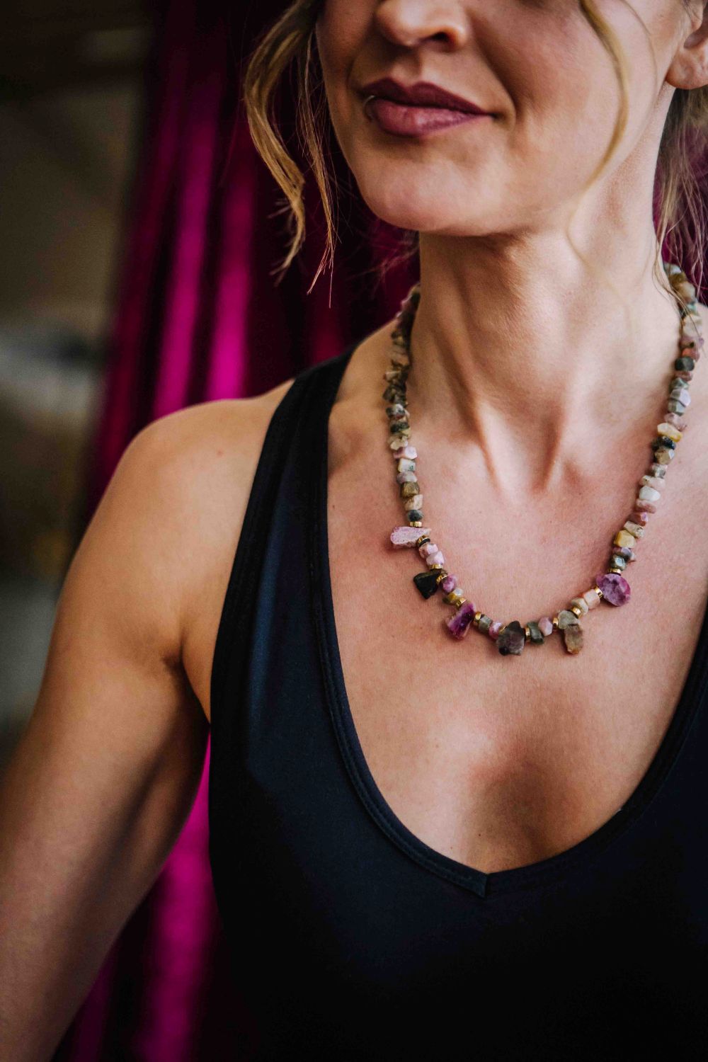 Woman wearing a black tank top and colorful beaded necklace with a blurred pink curtain background