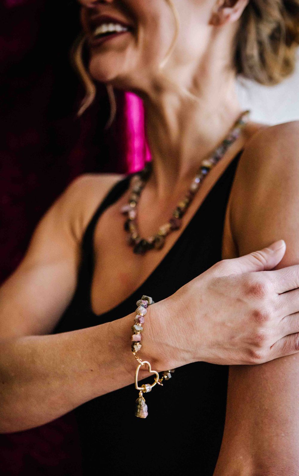 Woman wearing a gold bracelet with heart charm and pink beads, holding a pink object.